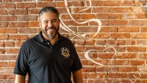 Farm Table Coffee House and Los Hermanos owner smiling in front of a brick wall.