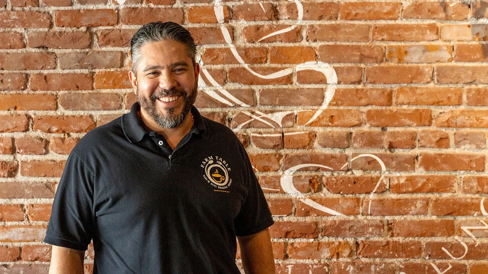 Farm Table Coffee House and Los Hermanos owner smiling in front of a brick wall.