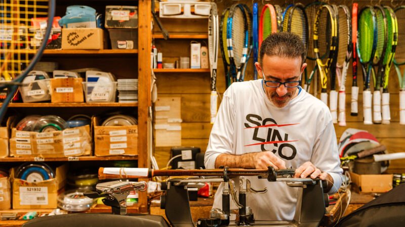 Owner of Westwood Sporting Goods stringing a tennis racket surrounded by rackets, spools, etc.