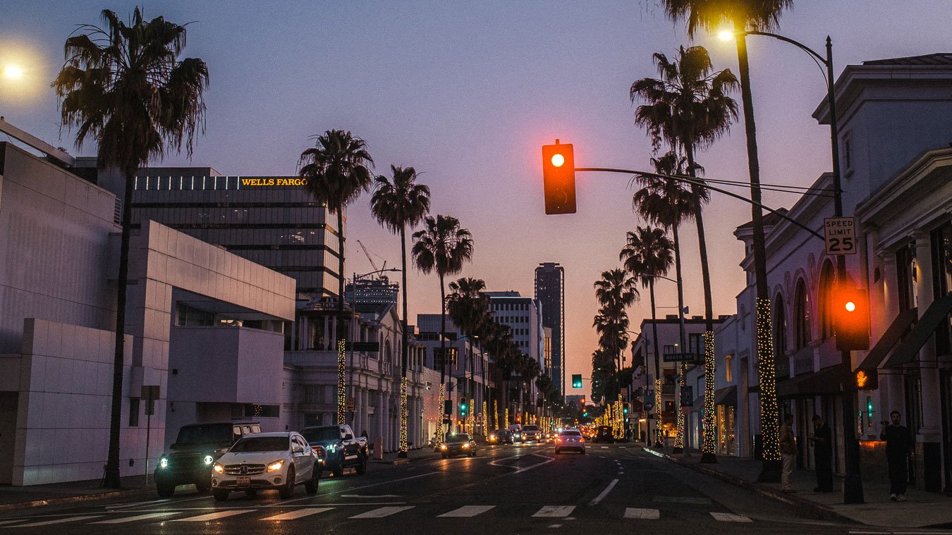 Dusk with street lights signaling red, a crosswalk, and buildings on a palm tree lined Wilshire Boulevard.