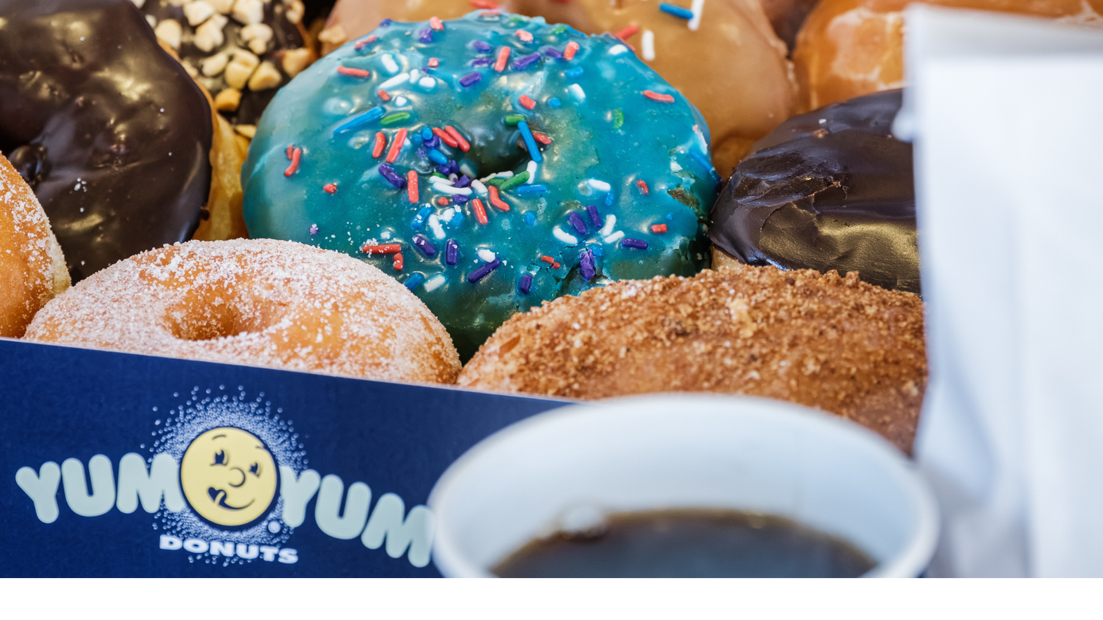 Yum Yum Donuts – box of assorted glazed, chocolate and sprinkle doughnuts next to a cup of coffee.