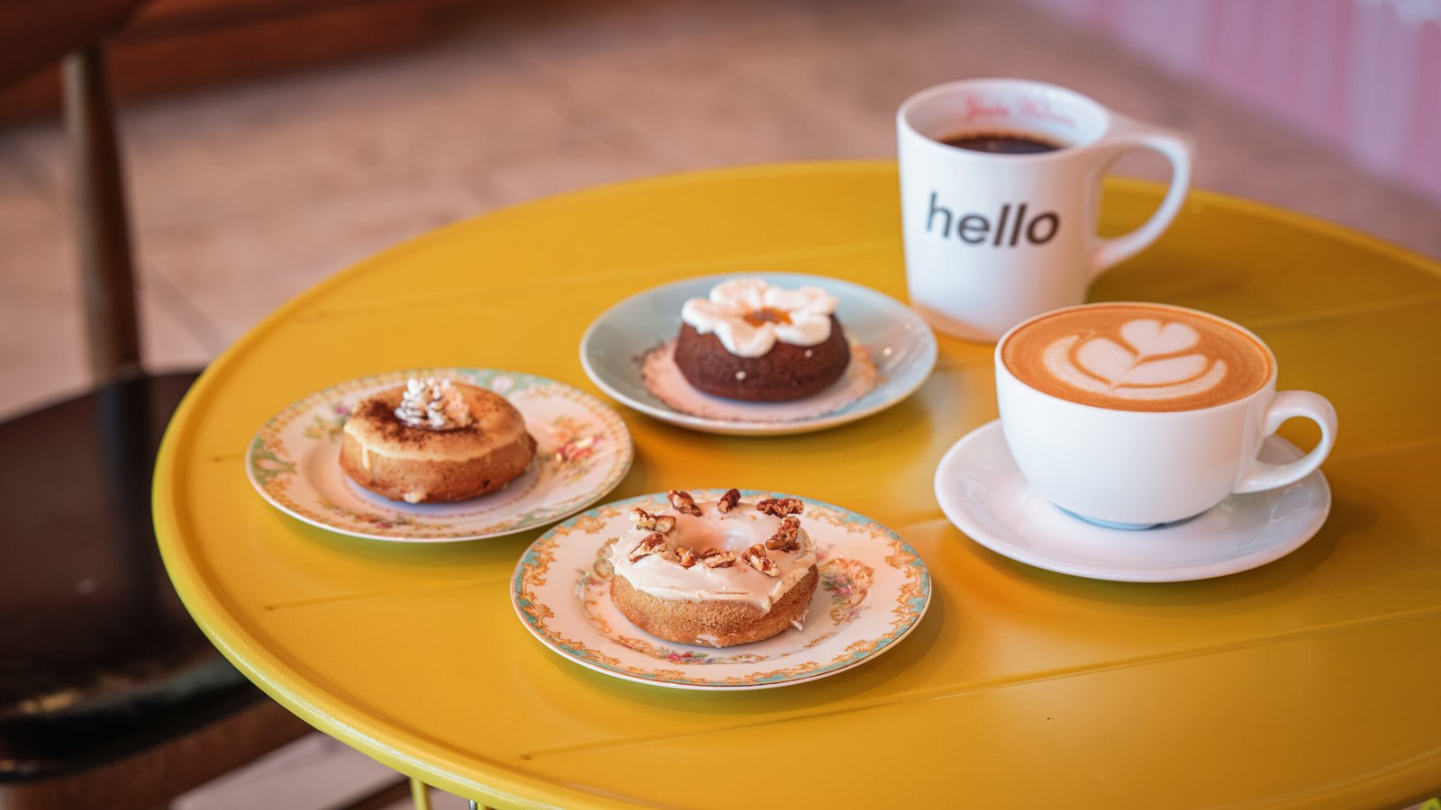Hello, You're Welcome – latte in a mug reading 'hello' beside plates of glazed and chocolate doughnuts on a yellow café table.