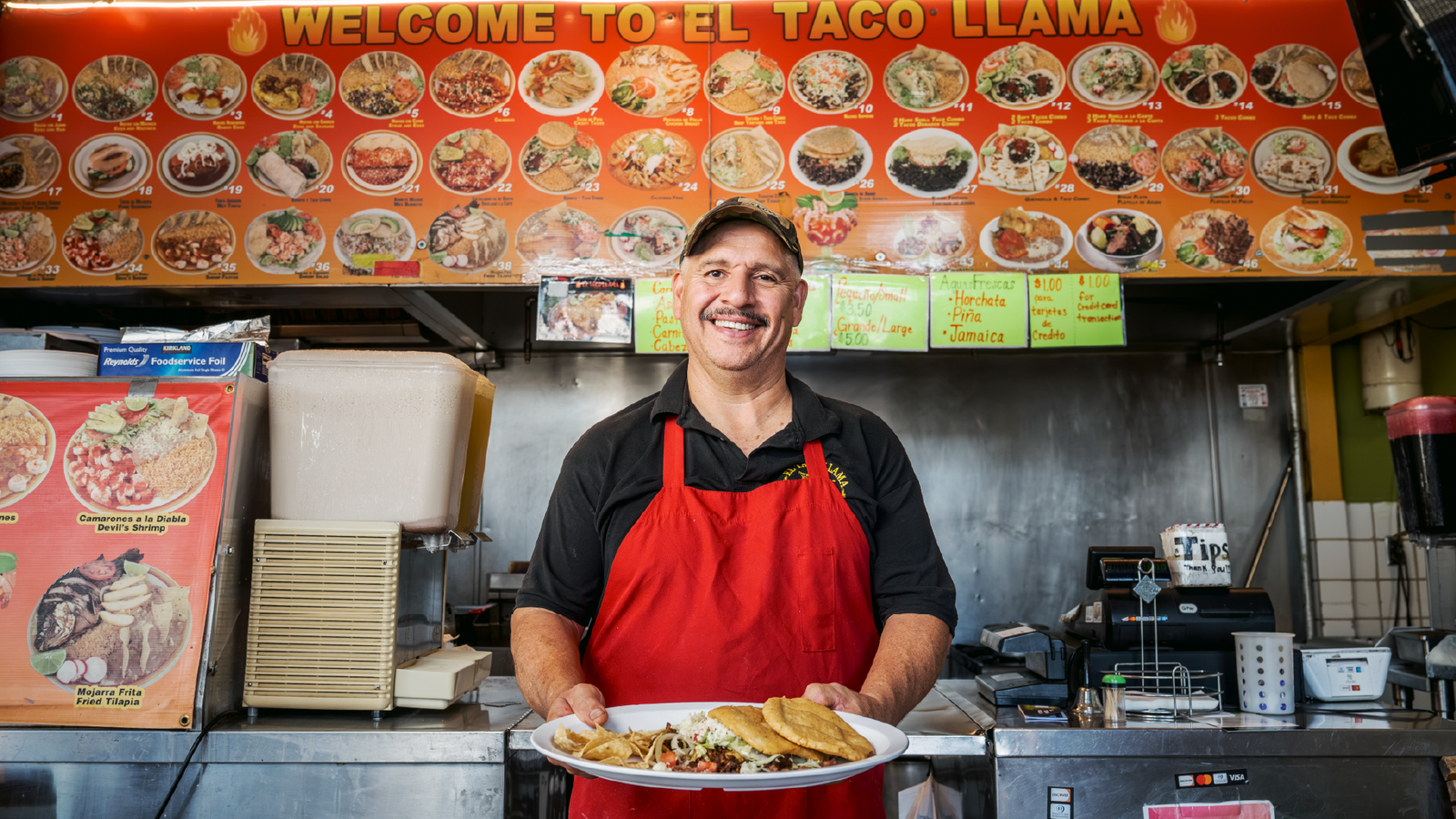 El Taco Llama – cook in a red apron holding a plate of tacos beneath the restaurant's photo menu board.