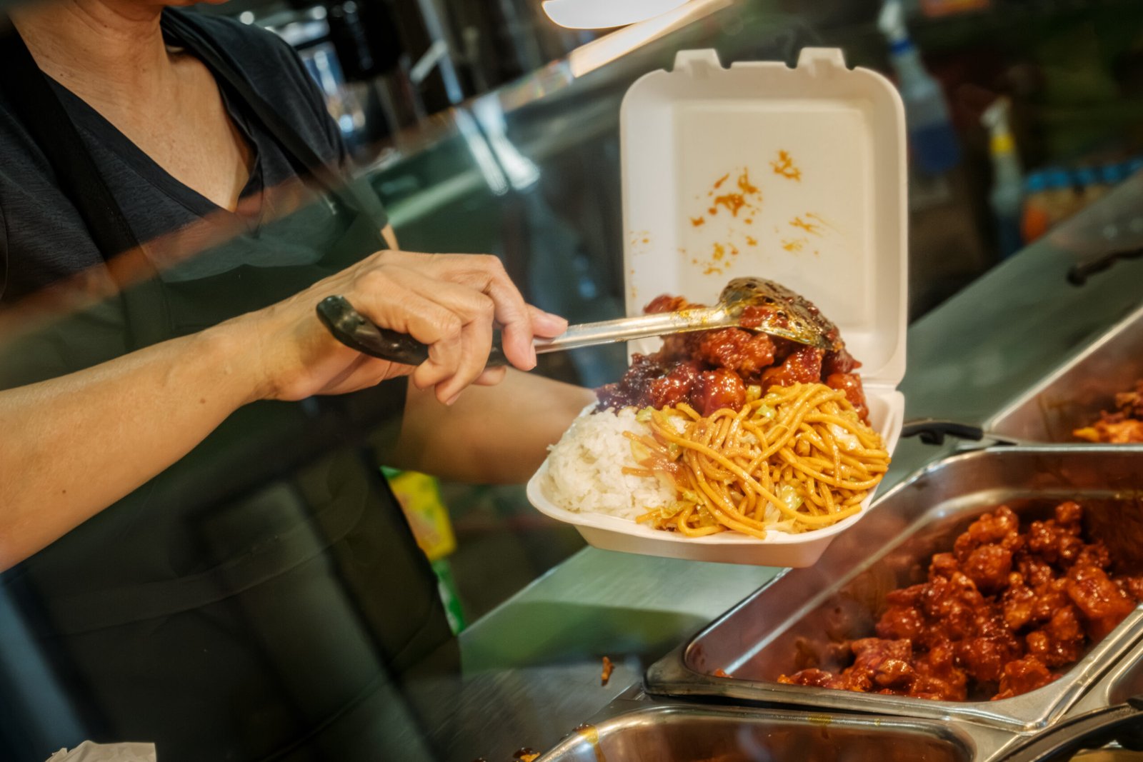 O K Chinese Restaurant – cook filling a takeout container with chow mein and orange chicken from the steam table.