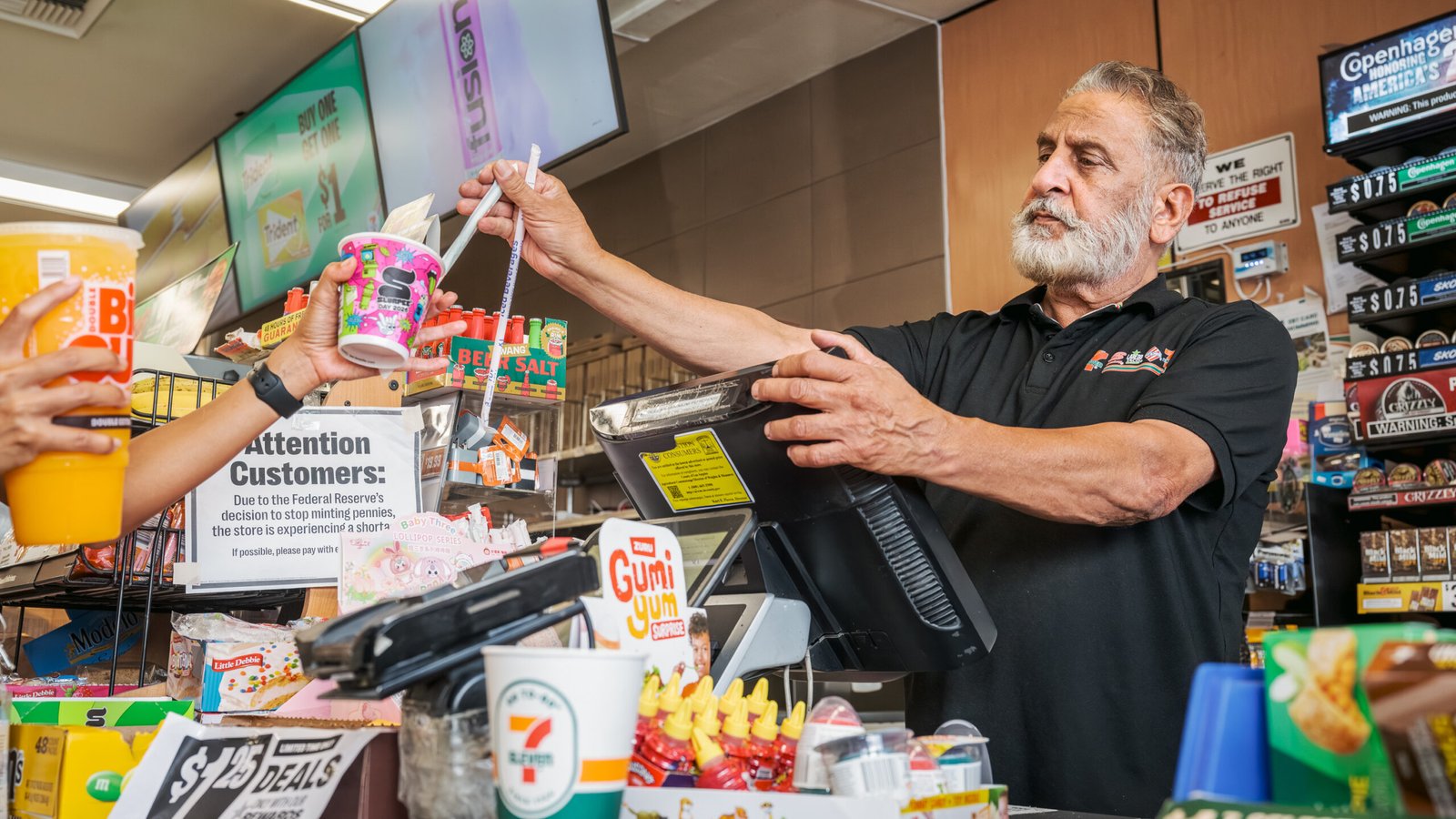 7-Eleven – store clerk bagging an order at the checkout counter surrounded by snacks, drinks and Slurpee cups.
